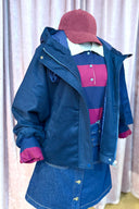 Close-up van een marineblauw jack gedragen over een gestreepte rugby-polo, een denim rokje en een bordeaux corduroy pet.