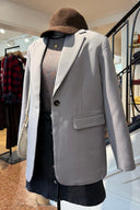 Close-up van een etalagepop bij Sienna met een herfstoutfit: een oversized lichtgrijze blazer gedragen over een donkere top met print en een subtiele ketting. De look is gestyled met een zwarte denim rok met knopen, een bruine corduroy pet en een lichte schoudertas.